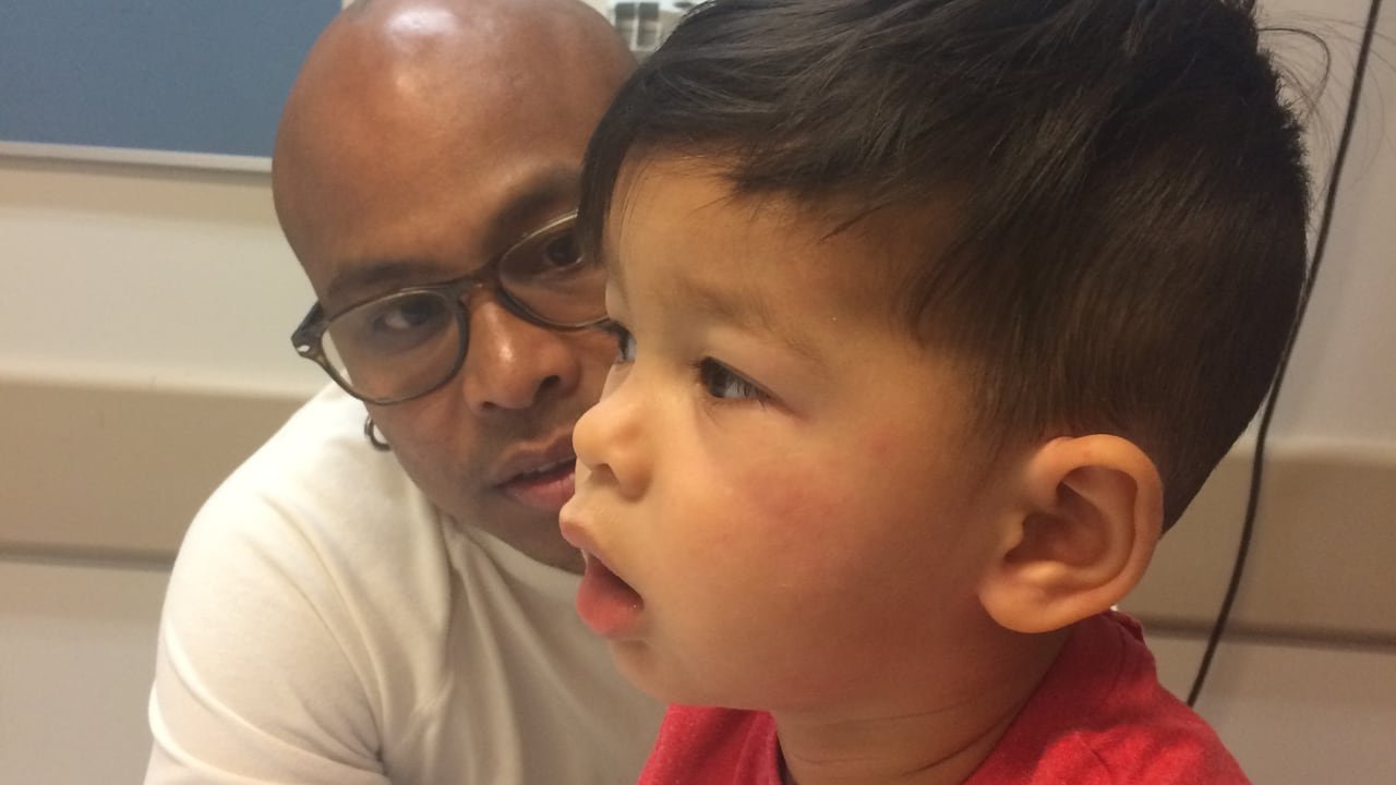 default-173 In a hospital room, a man looks concerned at a boy breathing with his mouth open and who has hives on his cheeks.
