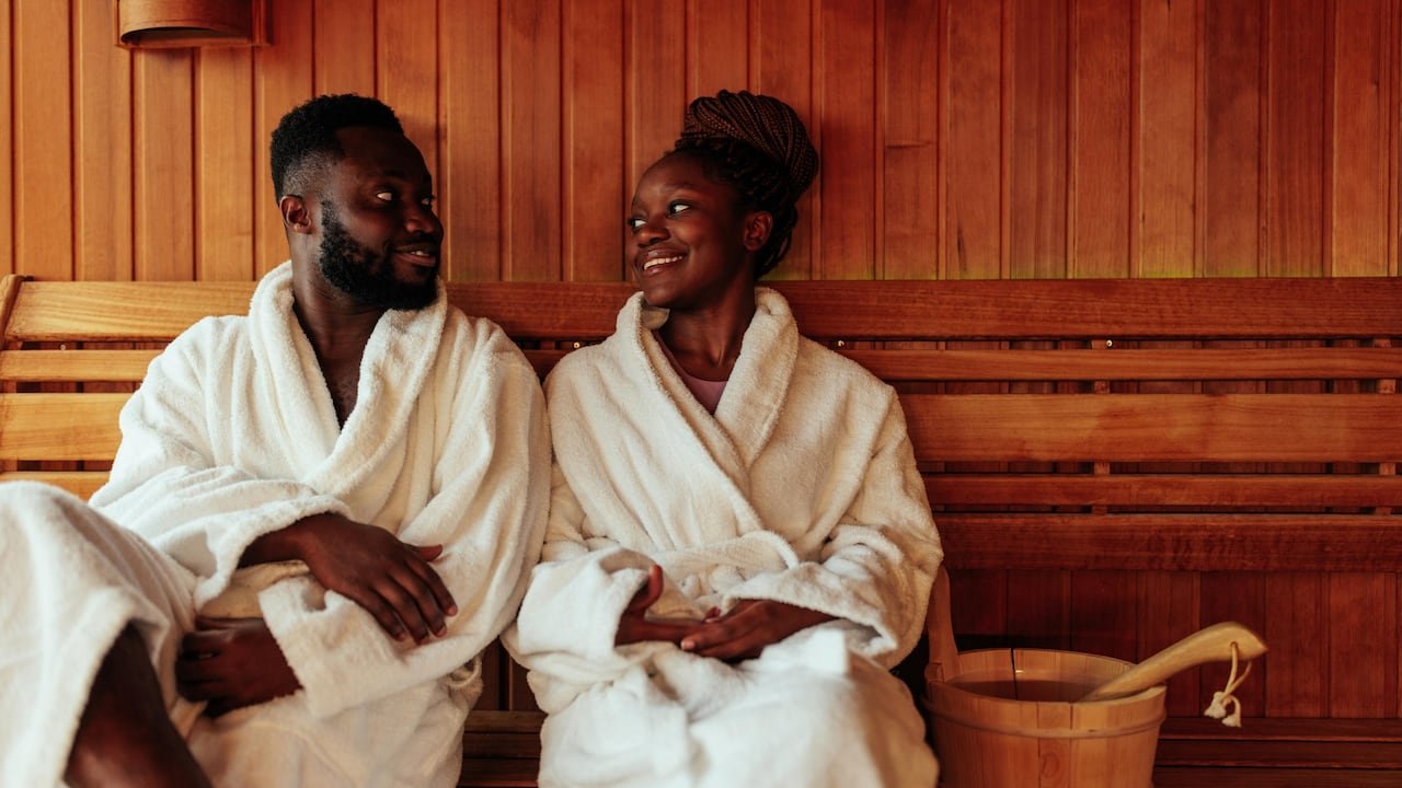 default-21 A couple in bath robes sitting in a wood-paneled sauna, smiling at each other.